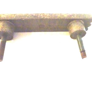 May include: A weathered, rectangular metal drawer pull with two cylindrical posts and threaded screws. The pull has decorative edges and a tarnished, aged appearance, suggesting vintage hardware.