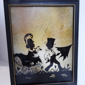May include: Framed silhouette art featuring two figures in a whimsical scene. The black silhouettes are set against a gold and beige background, with a black frame. The artwork depicts a man with an umbrella and a woman.