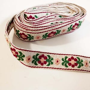 May include: Rolled-up cream-colored ribbon with a floral pattern. The ribbon features a repeating design of red and pink flowers with green leaves, bordered by a thin red line. The ribbon is likely used for crafts or decoration.