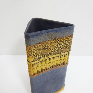 May include: A triangular ceramic vase with a textured surface. The vase features a blue base, a yellow middle section with a lace-like pattern, and a blue rim. The vase is approximately 8 inches tall.