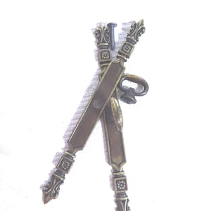 May include: Two antique brass cabinet pulls and a screw. The pulls have a rectangular design with ornate floral and scrollwork details. The pulls are crossed, with a small handle on one. The hardware is likely for a cabinet or drawer.