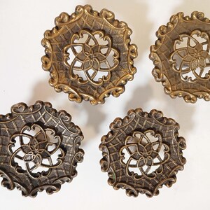May include: Four circular brass-toned buttons featuring an intricate openwork center design with a floral motif, surrounded by a textured grid pattern and a scalloped edge. Each button has a metallic finish with dark highlights in the crevices.