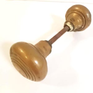 May include: An antique brass door knob with a rounded, ridged handle and a circular backplate. The knob has a warm, golden hue and shows signs of age, suggesting it is vintage. The connecting rod is a darker, contrasting color.