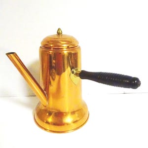 May include: A copper-colored coffee pot with a spout, lid, and a black wooden handle. The pot has a shiny finish and a small, gold-colored knob on the lid. The handle is attached with a gold-colored bracket.
