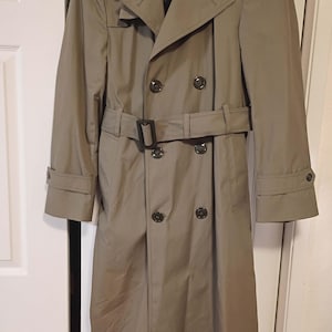 May include: A tan trench coat with a double-breasted design, featuring a belt at the waist and buttoned cuffs. The coat has a classic collar and multiple buttons. The coat is hanging on a door.
