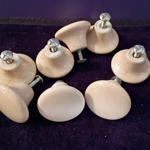 May include: A set of seven light beige ceramic cabinet knobs, each featuring a smooth, rounded top and a central metal screw attachment. The knobs are arranged in a cluster, showcasing their glossy finish and uniform, circular shape.