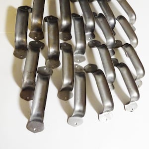 May include: A collection of vintage metal handles, likely for drawers or cabinets, arranged on a white surface. The handles are curved with a slightly aged, silver-gray finish, and each has mounting holes for attachment. The overall aesthetic suggests a focus on restoration or antique hardware.