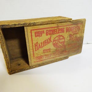 May include: A vintage wooden box with a sliding lid. The box is made of weathered wood and features red lettering that reads "Halifax Boneless Codfish". The box is open, revealing the interior.