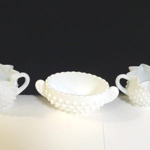 May include: Three white hobnail milk glass serving pieces. The set includes two handled creamers and a round bowl with handles. The pieces have a ruffled edge and a textured surface.