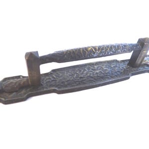 May include: Bronze-toned door pull with a textured, organic design. The handle is supported by two uprights and is mounted on a rectangular backplate. The surface has a hammered appearance.