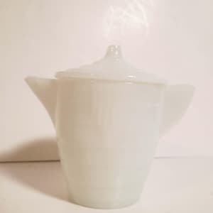 May include: A white, opaque, vintage-style creamer with a matching lid. The creamer has a spout and a handle, and the body features horizontal ribbed detailing. The lid has a small, rounded knob.