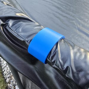 Może przedstawiać: Jasnoniebieski, plastikowy klips w kształcie litery C zamocowany wokół czarnej, wyściełanej krawędzi zabezpieczającej trampoliny. Klips ma gładką, solidną powierzchnię i służy do mocnego utrzymywania czarnego materiału winylowego na ramie.
