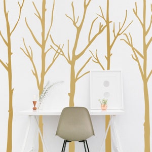 May include: A minimalist home office setup featuring a white desk, a gray chair, and a framed artwork. The wall is adorned with gold tree decals. A small vase with flowers and a candle holder add decorative touches.