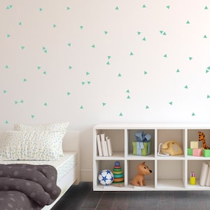 Puede incluir: Un dormitorio infantil con una cama blanca, una estantería blanca y una pared decorada con calcomanías triangulares turquesas. La estantería contiene juguetes, libros y objetos decorativos. La cama tiene una sábana y almohada estampadas, y una manta gris.