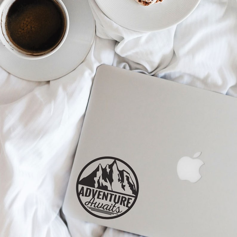 May include: A silver laptop with a black circular sticker that reads "ADVENTURE AWAITS" with a mountain graphic. A cup of coffee and a plate with a pastry are in the background on a white sheet.