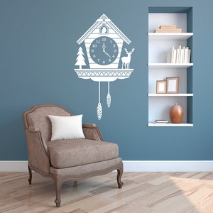 Puede incluir: Un vinilo blanco de pared de un reloj de cuco con diseño de pájaro, ciervo y pino. La esfera del reloj muestra la hora 12:05. La calcomanía está montada en una pared azul sobre un sillón marrón con un cojín blanco.