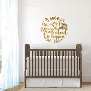 May include: A brown wooden crib with a white mattress and a white skirt. Above the crib, a gold-colored wall decal displays the quote, "As soon as I saw you, I knew a grand adventure was about to happen. - AA Milne."