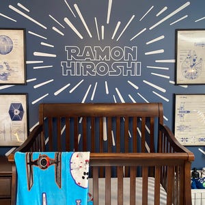 May include: A dark blue nursery with a white starburst design on the wall. The name "RAMON HIROSHI" is written in white letters on the wall. A crib with a wooden frame is in the center of the room. A blue blanket with a Star Wars spaceship design is on the crib. There are three framed prints on the wall with Star Wars spaceship designs.