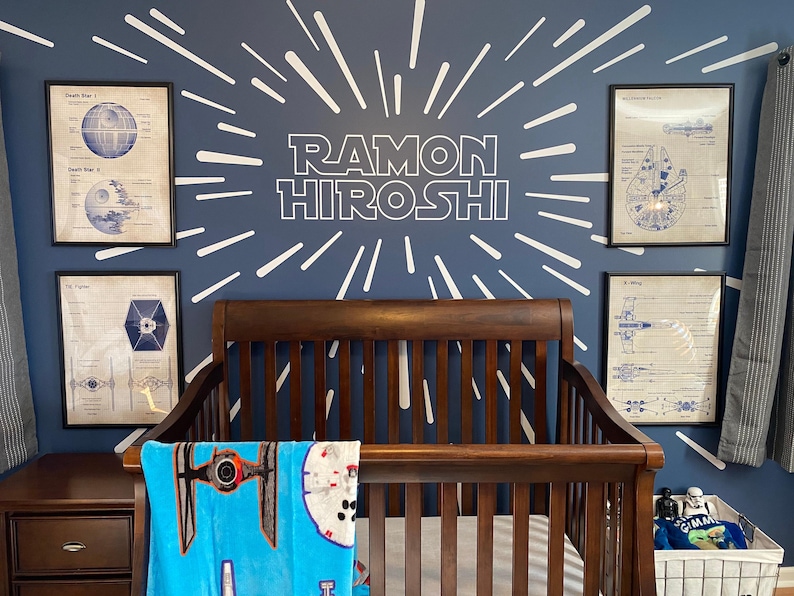May include: A dark blue nursery with a white starburst design on the wall. The name "RAMON HIROSHI" is written in white letters on the wall. A crib with a wooden frame is in the center of the room. A blue blanket with a Star Wars spaceship design is on the crib. There are three framed prints on the wall with Star Wars spaceship designs.