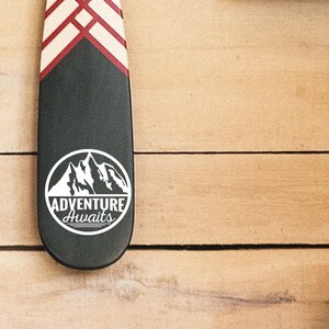 May include: A black paddle with a white mountain graphic and the words "ADVENTURE AWAITS" in a white circle. The top of the paddle has a red and white geometric design. The paddle is on a wooden surface.