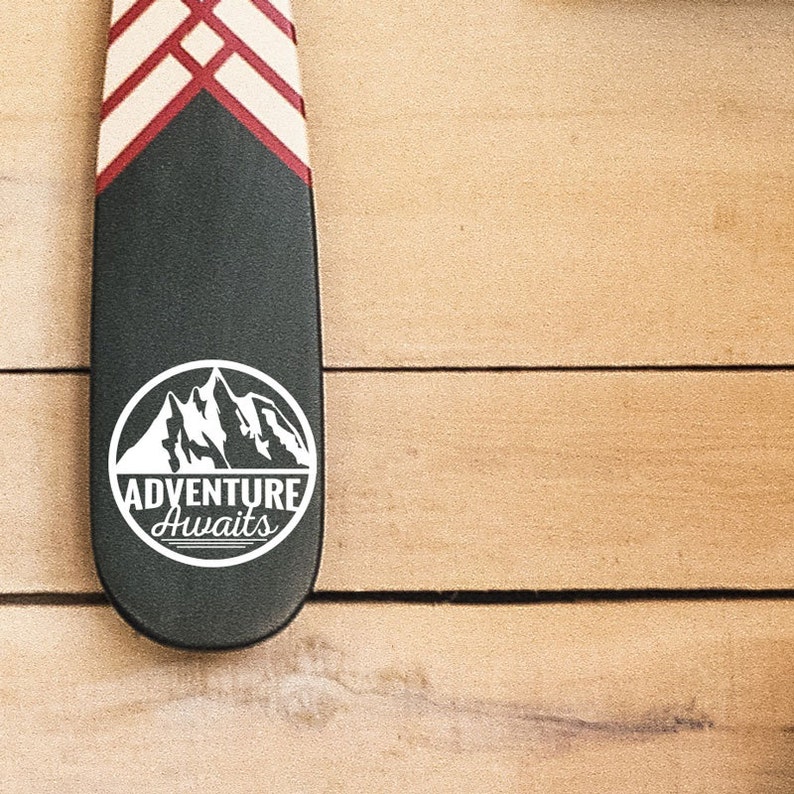 May include: A black paddle with a white mountain graphic and the words "ADVENTURE AWAITS" in a white circle. The top of the paddle has a red and white geometric design. The paddle is on a wooden surface.