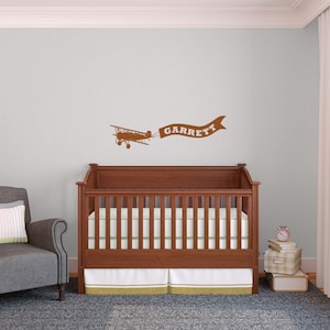 May include: A brown wooden crib with a mattress and bedding, a gray armchair, and a lamp. A brown airplane decal with a banner that reads "GARRETT" is on the wall. The room has a light blue wall color and a gray carpet.
