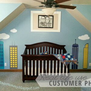 Puede incluir: Una habitaci&oacute;n de beb&eacute; con un esquema de color azul y blanco. La habitaci&oacute;n cuenta con una cuna con una colcha azul y roja, un ventilador de techo y un marco de fotos con una foto de una pareja. Las paredes est&aacute;n decoradas con un mural de ciudad con nubes blancas y edificios amarillos, azules y grises.
