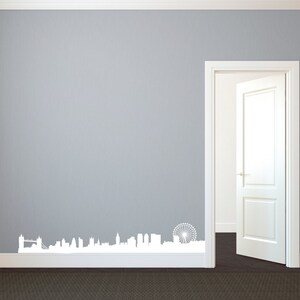 May include: A white vinyl wall decal of the London skyline, including the Tower Bridge and the London Eye, is applied to a gray wall. The decal is placed near the floor, above a dark brown carpet. A white door is open to the right.