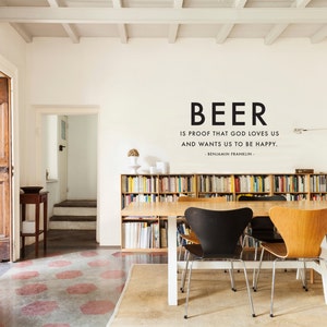 May include: An interior scene with a wooden table and chairs. The wall displays the quote "BEER is proof that God loves us and wants us to be happy." A bookshelf is filled with books. The floor has a patterned design.