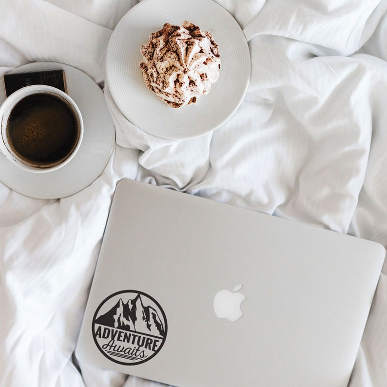 May include: A laptop with a black and white "Adventure Awaits" sticker, a cup of coffee, a dessert, and a chocolate bar on a white bedspread. The laptop is silver and has the Apple logo.