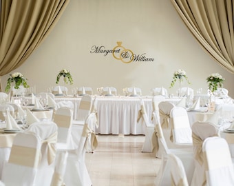 May include: A wedding reception setting with tables set for guests. White tablecloths and chair covers are accented with beige ribbons. Floral arrangements and elegant glassware adorn the tables. The wall features the names "Margaret & William" with wedding rings.