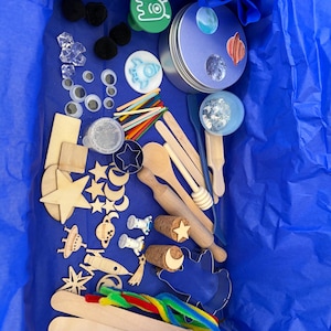 May include: A space-themed craft kit containing a round metal tin, wooden stars, moons, rockets, and astronaut figures. Includes googly eyes, clear plastic gems, colorful pipe cleaners, wooden spoons, a rolling pin, and craft sticks in a blue box.