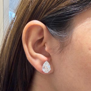 May include: A pear-shaped stud earring featuring a clear, faceted stone set in a silver-toned metal prong setting. The earring is positioned on the earlobe, showcasing the reflective quality of the stone and the secure metal mounting structure.