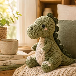 Puede incluir: Un peluche de dinosaurio tejido a ganchillo en color verde oliva con una barriga color crema y pequeñas mejillas rosadas. El juguete presenta una hilera de púas redondeadas a lo largo de la espalda y una cola larga. Tiene ojos de botón negros.