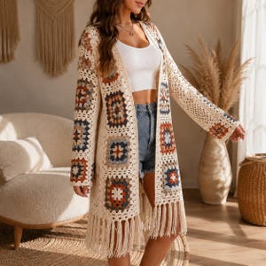 May include: A long-sleeved crochet cardigan featuring a cream-colored base with a multi-colored granny square pattern in shades of rust, navy, and grey. The garment has an open front, a straight hemline, and long fringe trim along the bottom edge.
