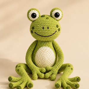 May include: A handmade crochet frog toy in shades of light and dark green with a cream-coloured belly. The frog features large black plastic eyes with white borders, a stitched dark brown smile, and textured spots on its head and legs.