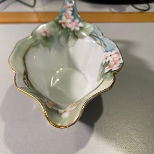 May include: A white porcelain gravy boat with a scalloped rim and gold trim. The exterior features a hand-painted design of soft pink flowers and green leaves against a pale blue and white background, with a pointed spout and a curved handle.