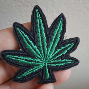 以下が含まれることがあります： 7枚の葉を持つ葉の形をした刺繍ワッペン。鮮やかな緑色の糸で細部が表現されており、無地の黒い背景に、葉の形全体を縁取る厚い黒いステッチのボーダーが施されています。