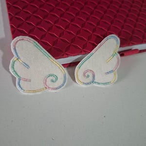 Op de afbeelding: Twee witte vilt patches in de vorm van vleugels met geborduurde randen in pastel regenboogkleuren, waaronder roze, geel, blauw en groen. Het borduurwerk volgt de gebogen randen en vormt een spiraalvormig detail aan de basis.