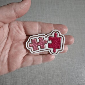 May include: An embroidered patch featuring two interlocking puzzle pieces in shades of pink. The left piece is a lighter rose pink, while the right piece is a deeper magenta. Both are outlined in brown thread against a white fabric border.