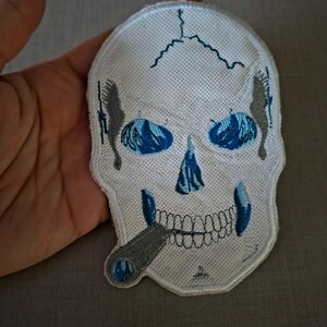 May include: An embroidered patch shaped like a human skull in white thread with blue accents in the eye sockets and nasal cavity. The skull features a grey cigar in its mouth and grey shading along the sides, with a stitched border edge.