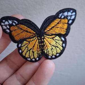 May include: An embroidered butterfly patch featuring vibrant orange and yellow wings with detailed black veining and a thick black border. The wing tips are accented with small white spots, creating a realistic monarch butterfly design.
