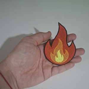 Könnte beinhalten: Ein gestickter Aufnäher in Flammenform mit schwarzem Rand und einem Farbverlauf, der von einem hellgelben Zentrum über einen orangefarbenen Mittelteil bis zu einer dunkelroten Außenschicht reicht und ein stilisiertes Feuermotiv bildet.