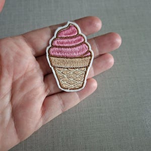 Op de afbeelding: Een geborduurde patch in de vorm van een softijsje in een hoorntje. Het bovenste gedeelte heeft roze draad in een wervelpatroon, terwijl het onderste hoorntje beige is met een gekruiste wafelstructuur. De hele patch heeft een witte rand.