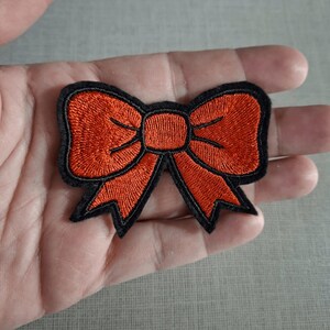 Op de afbeelding: Een geborduurde patch in de vorm van een strik, voorzien van feloranje draad voor de lussen en uiteinden van de strik, omlijnd met een dikke zwarte rand. Het ontwerp heeft een getextureerd oppervlak door het dichte borduurwerk.