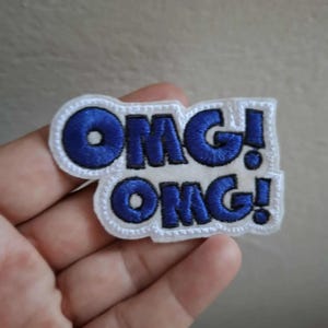 Op de afbeelding: Een geborduurde patch met de tekst OMG! OMG! in dikke blauwe letters met een dikke zwarte omlijning. De letters staan op een witte achtergrond met een witte rand die de contouren van de tekst volgt voor een gelaagd effect.