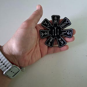 May include: A black, star-shaped embroidered patch featuring white stitched symbols in each of its eight points and a central design. The patch has a distinct white border outlining its geometric shape, with various unique markings embroidered in white thread.