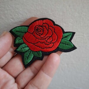 Puede incluir: Un parche bordado que presenta una rosa roja vibrante con costuras detalladas en rojo oscuro para dar profundidad. La rosa está rodeada de hojas verdes con textura de hilo visible, todo sobre un fondo de tela negra con un borde recortado.