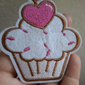 Op de afbeelding: Een cupcake-vormige geborduurde patch met een witte glazuurlaag, kleine roze spikkels en een bruine omlijning. Bovenop het glazuur zit een roze hartvormige kers met een bruin steeltje. De onderkant van de cupcake is wit met bruine verticale lijnen.