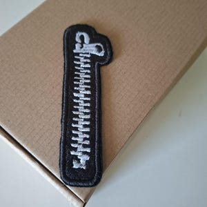 Può includere: Una toppa ricamata nera a forma di cerniera chiusa. La toppa presenta uno sfondo nero con dettagli in filo bianco che imitano i denti e il cursore di una cerniera, rifinita con un bordo cucito nero lungo tutto il perimetro esterno.